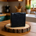  "BY RICEL Onyx Soul handcrafted charcoal soap bar standing upright on a rustic wood slice in a warm kitchen setting"