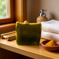 "BY RICEL Terra Bar travel size artisan soap bar on a wood bathroom shelf beside a clay bowl of turmeric powder, bamboo sticks, and folded white towels with warm window light"