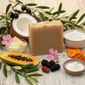 BY RICEL Orchard Bounty artisan soap bar surrounded by its key ingredients — olive oil, coconut, shea butter, rose geranium flowers, goat milk, carrot, green papaya, and mulberries — on a warm wooden surface