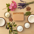 BY RICEL Sedona Earth artisan soap bar surrounded by its key ingredients — rose geranium flowers, lavender sprigs, cedarwood chips, olive oil, coconut, shea butter, goat milk powder, and kaolin clay — on a warm wooden surface